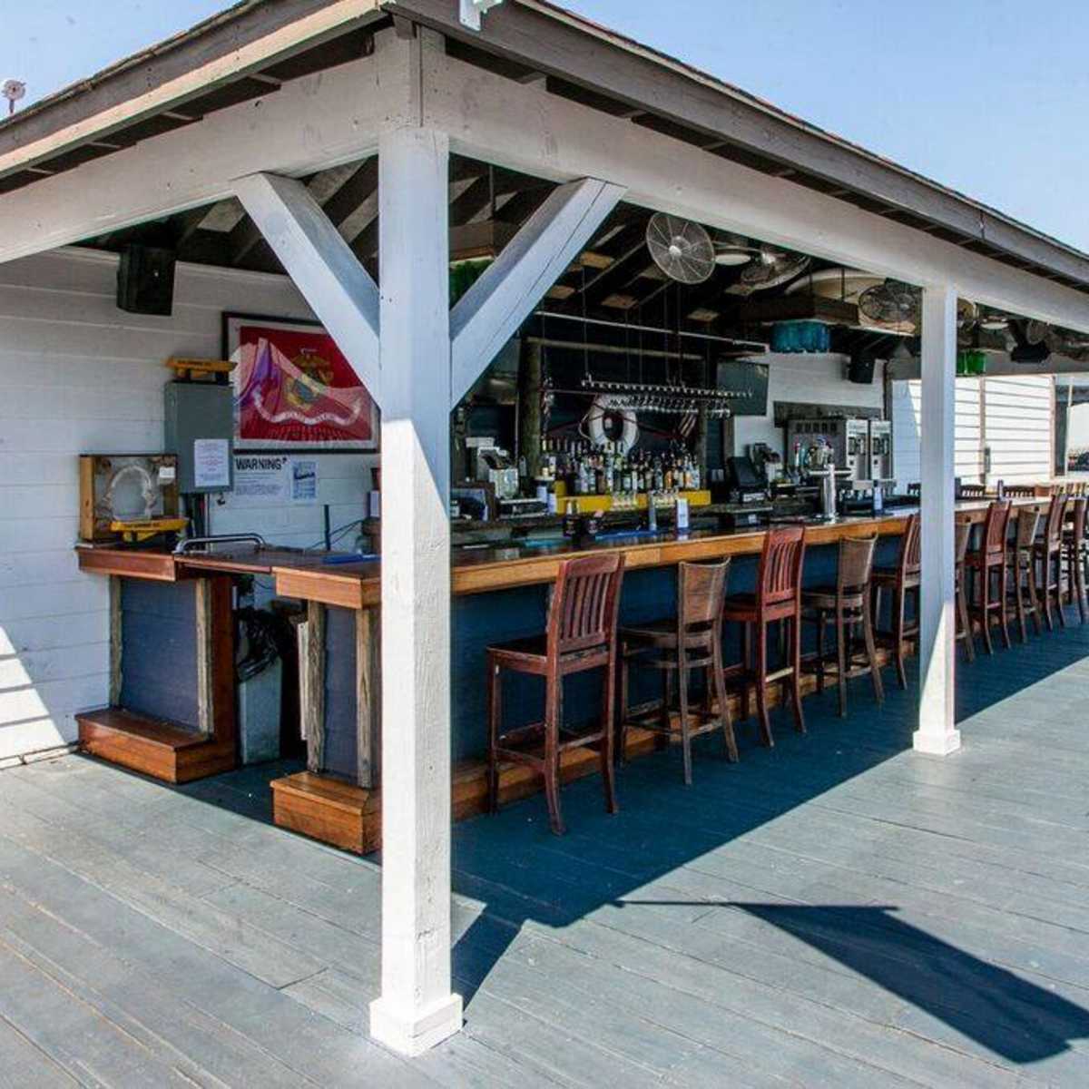 An outdoor bar with a wooden counter and high chairs stands under a roof supported by white posts, with a small American flag visible in the background.