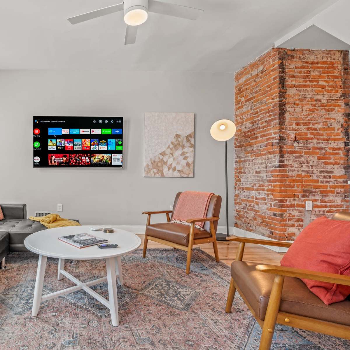 The living room features a modern television mounted on the wall above a light-colored coffee table, flanked by a dark couch and an armchair, with a brick accent wall in the background.