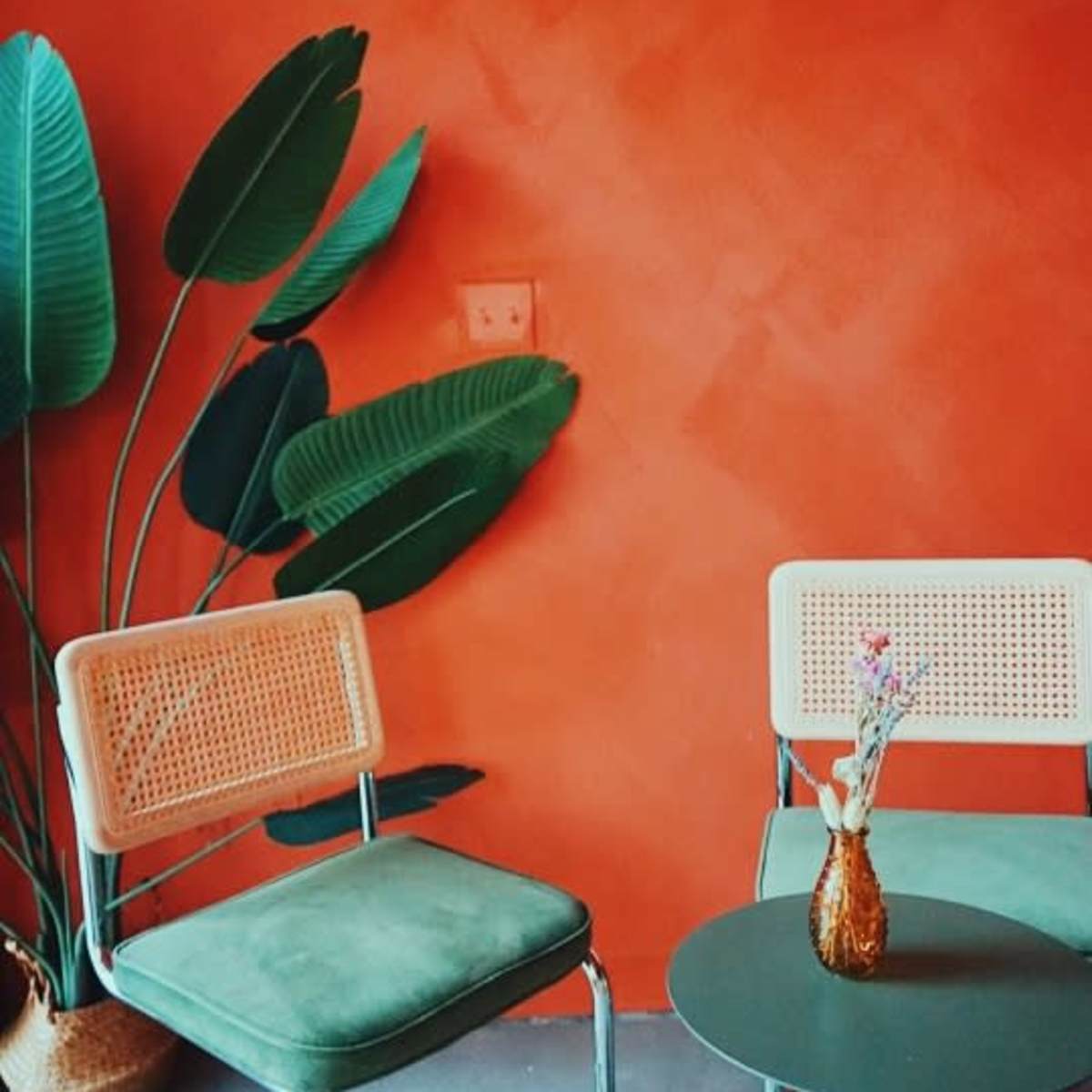 Two chairs beside a green table with a vase containing flowers against an orange wall decorated with large green leaves.