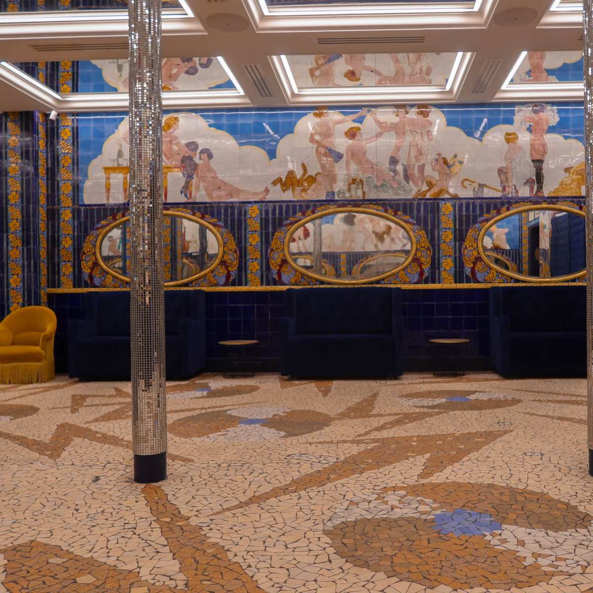 The interior features a mosaic tiled floor, ornate blue and gold wall decorations, and several mirrors reflecting an artistic mural.