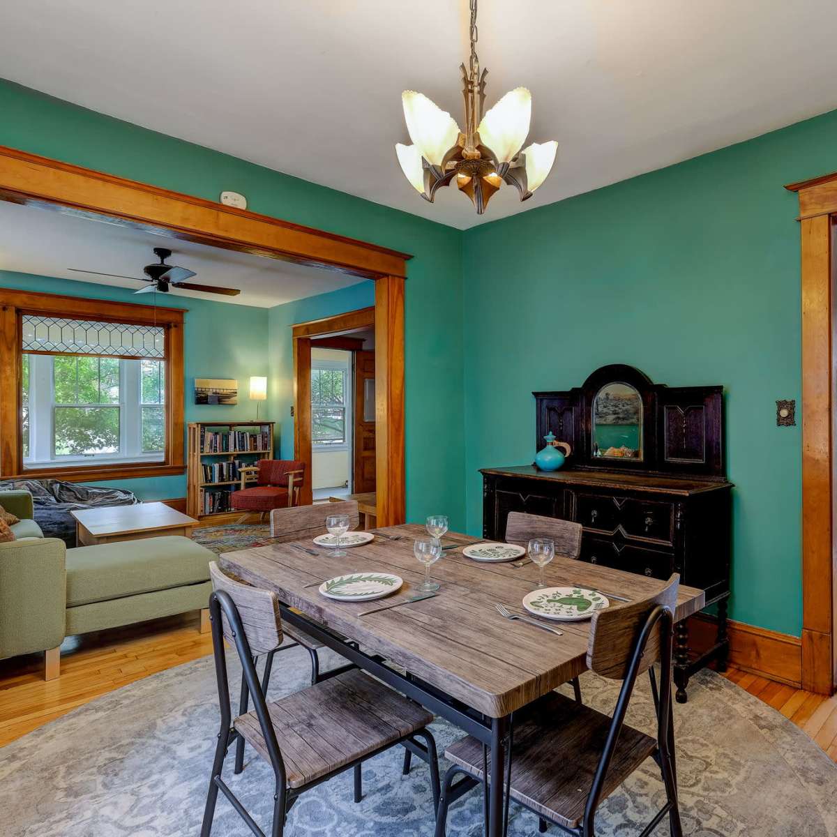 A dining area with a wooden table set for four, flanked by doorways leading to a green living room and a light-filled kitchen.