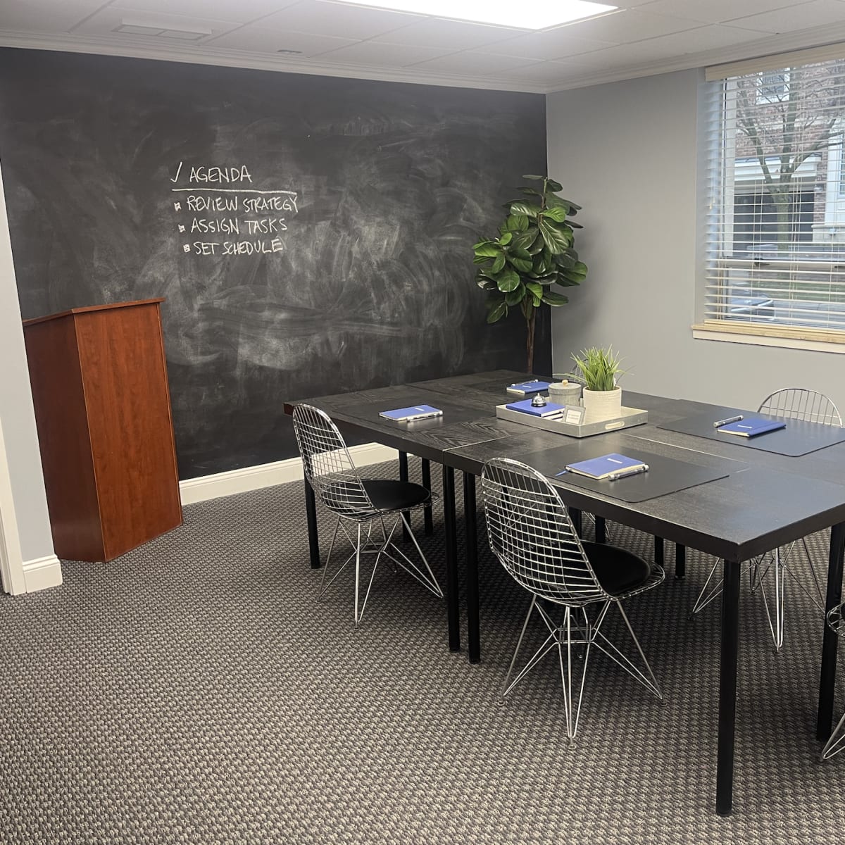 A conference room with a large black table surrounded by wire chairs, a chalkboard wall listing an agenda, and a bookshelf in one corner.