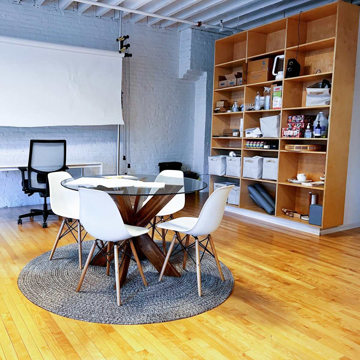 A bright office space features a round table with white chairs, a desk, a backdrop support, and shelves filled with various supplies.