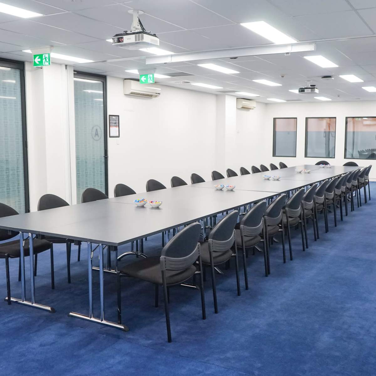 A long, gray conference table surrounded by black chairs is set up in a well-lit meeting room with large windows and blue carpeting.