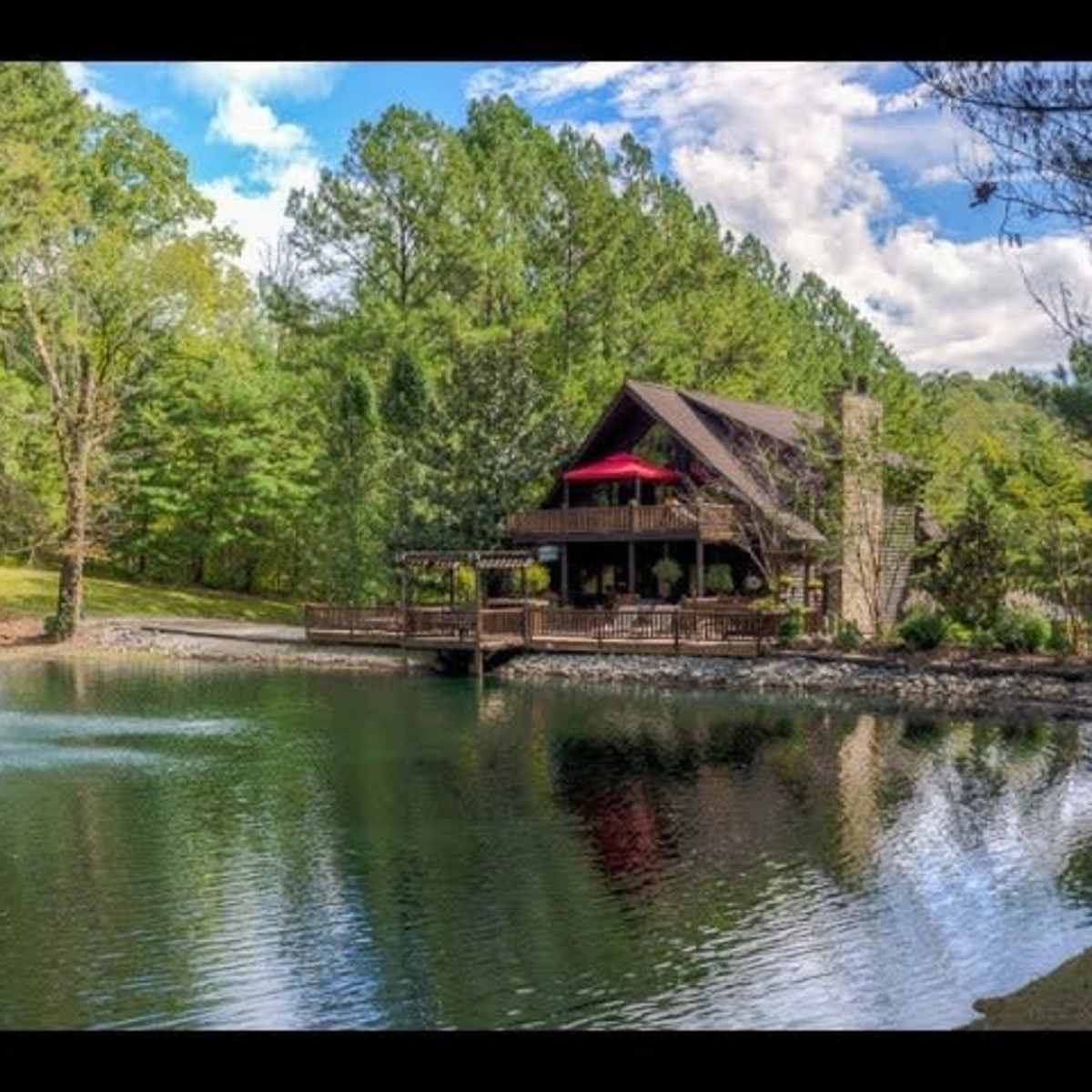 A two-story house with a large deck overlooks a pond featuring a fountain, surrounded by trees and a clear blue sky.