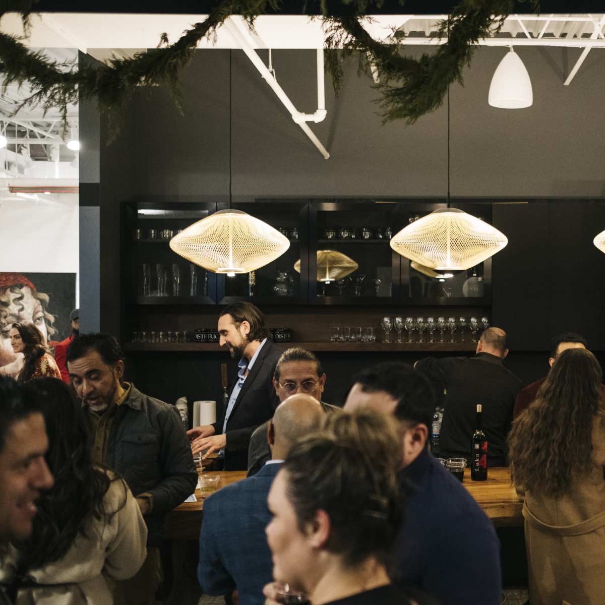 A crowded bar area features people conversing and a woman drinking from a glass, with modern pendant lights hanging above a long wooden counter.