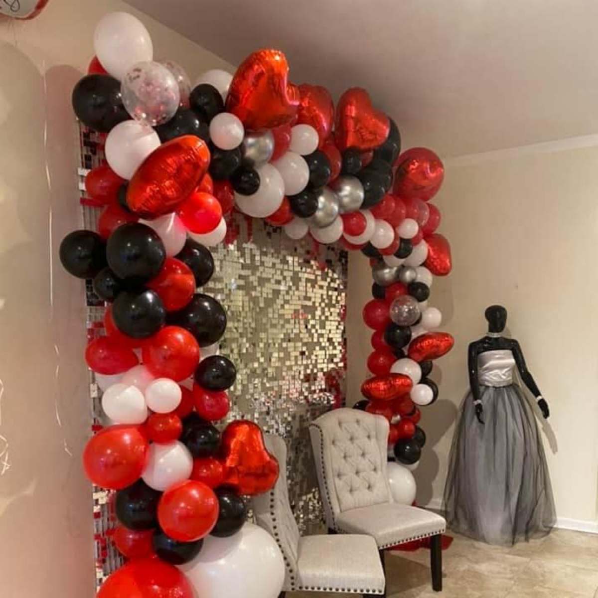 A decorative arch made of black, white, red, and silver balloons, framing a mirrored backdrop with two upholstered chairs in front and a mannequin dressed in a gray gown beside it.