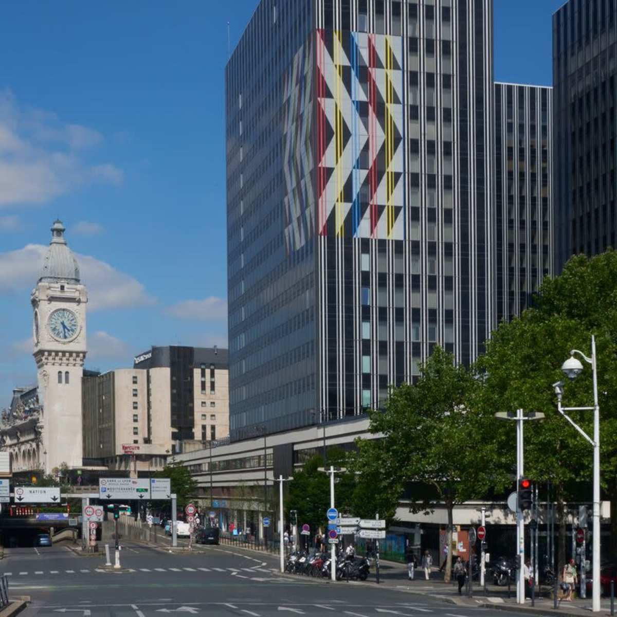 A modern multi-story building with a colorful geometric design on its facade, adjacent to a historic clock tower and a busy urban street.