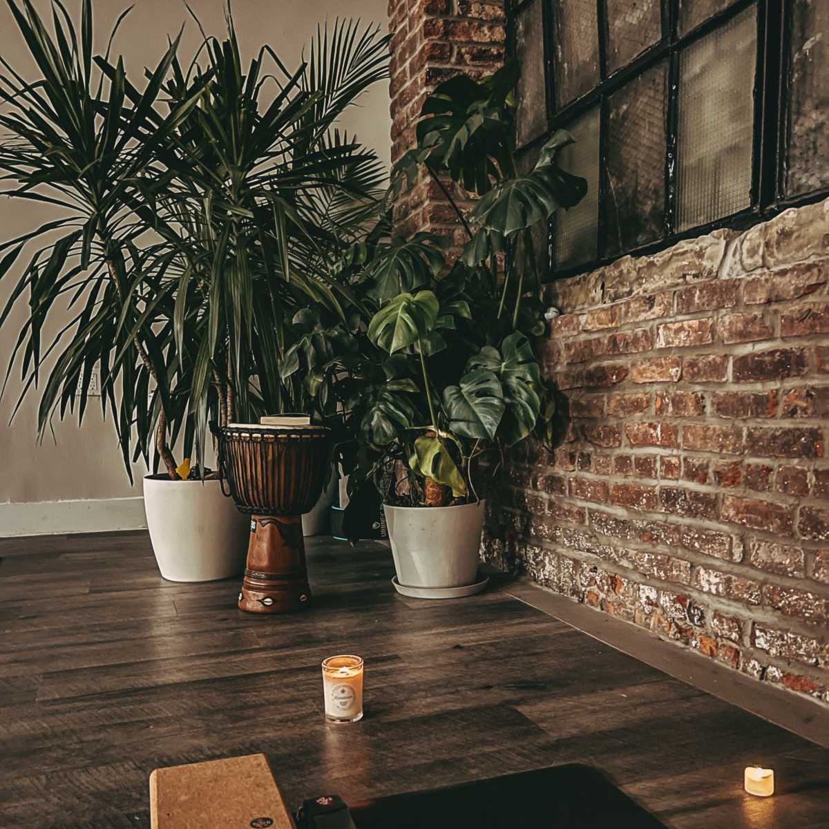 A yoga mat is placed on a wooden floor in front of potted plants and a candle near a large window with a brick wall backdrop.