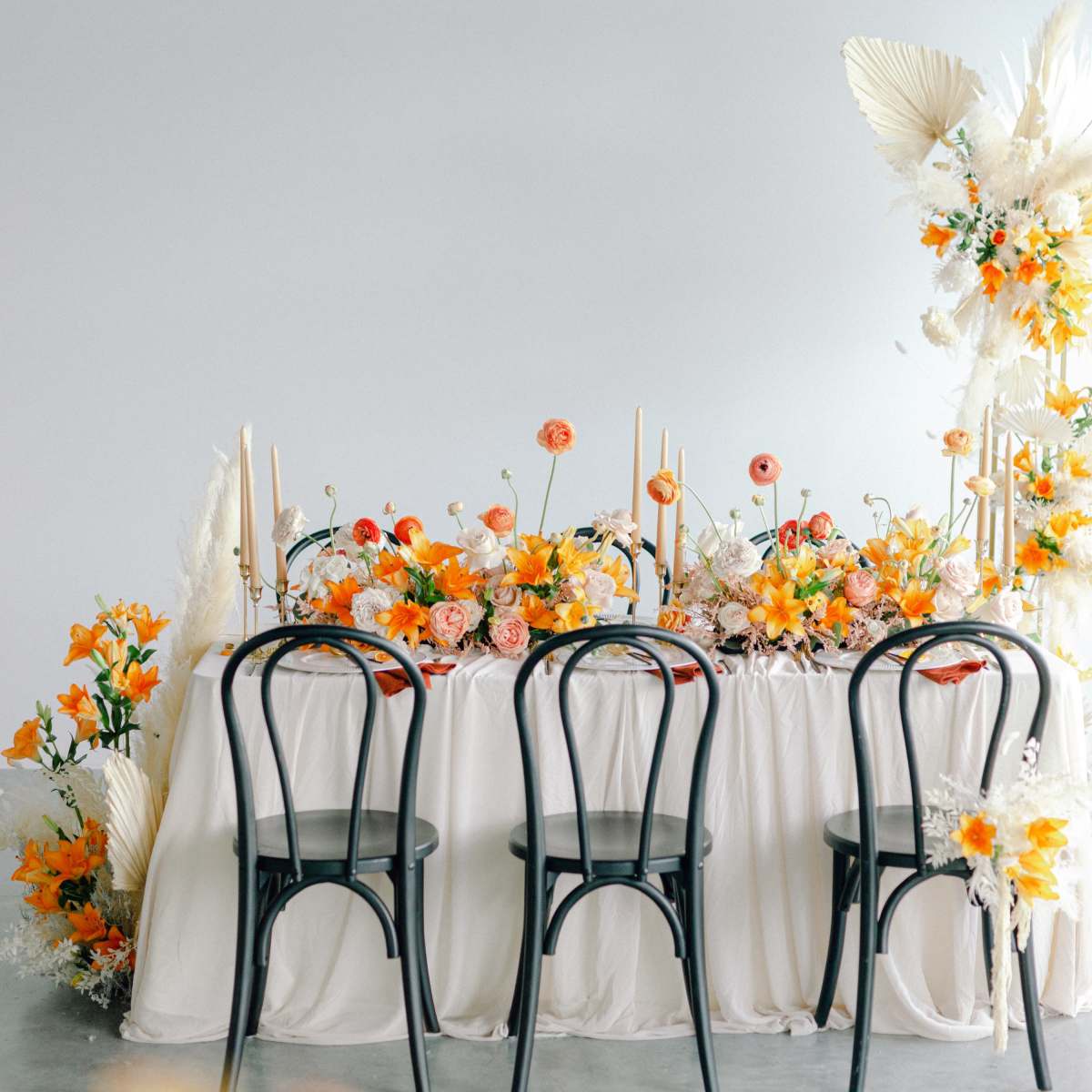 A decorated table with floral arrangements in yellow and orange hues is set with three black chairs facing it.