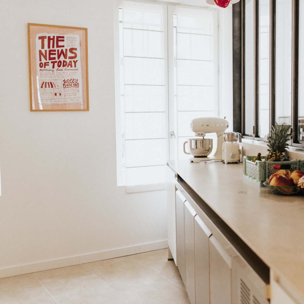 A modern kitchen features a countertop with a stand mixer, a basket of fruits, and a decorative poster on the wall, with sunlight streaming through the windows.