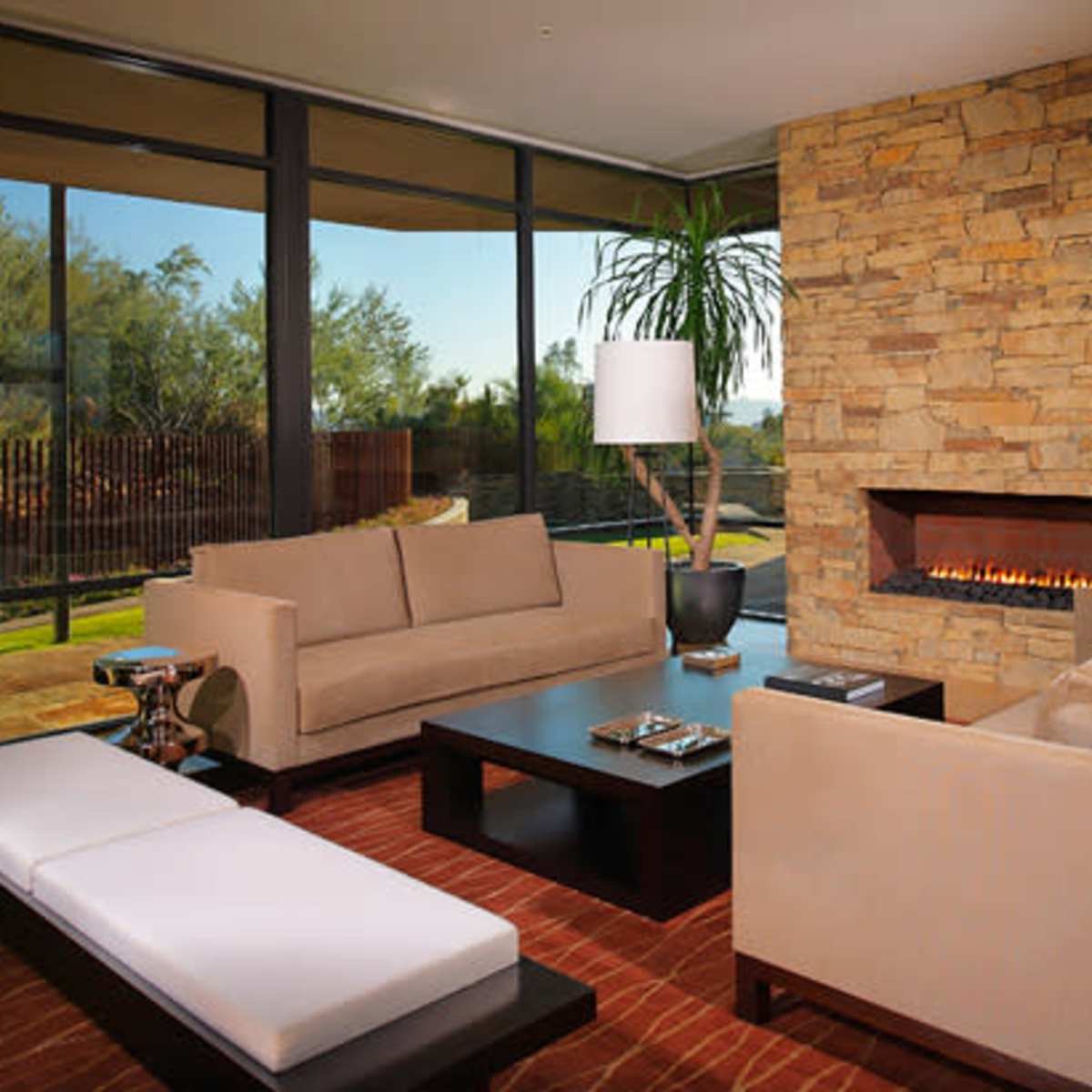 A modern living room features a stone fireplace, large windows, and contemporary furniture arranged around a coffee table.