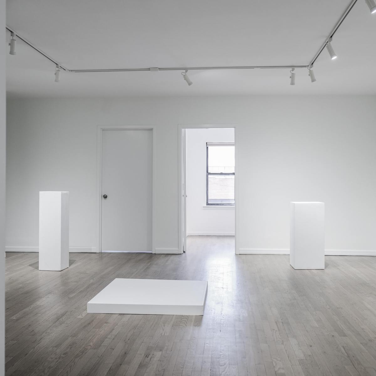 A minimalist art gallery with white walls, wooden floors, and three white pedestals arranged within the space.