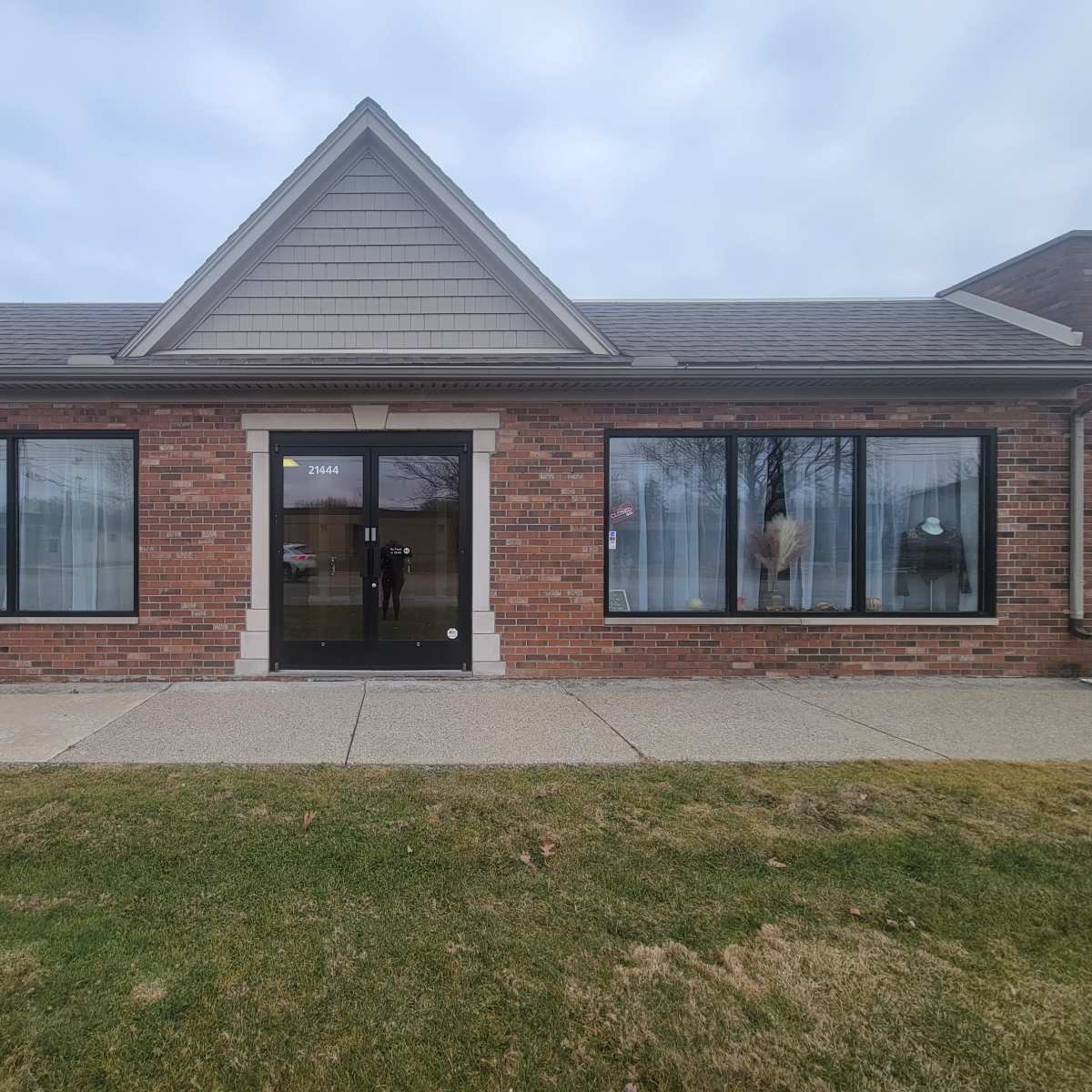 A brick building with large windows and a front door, set against a grassy area and cloudy sky.