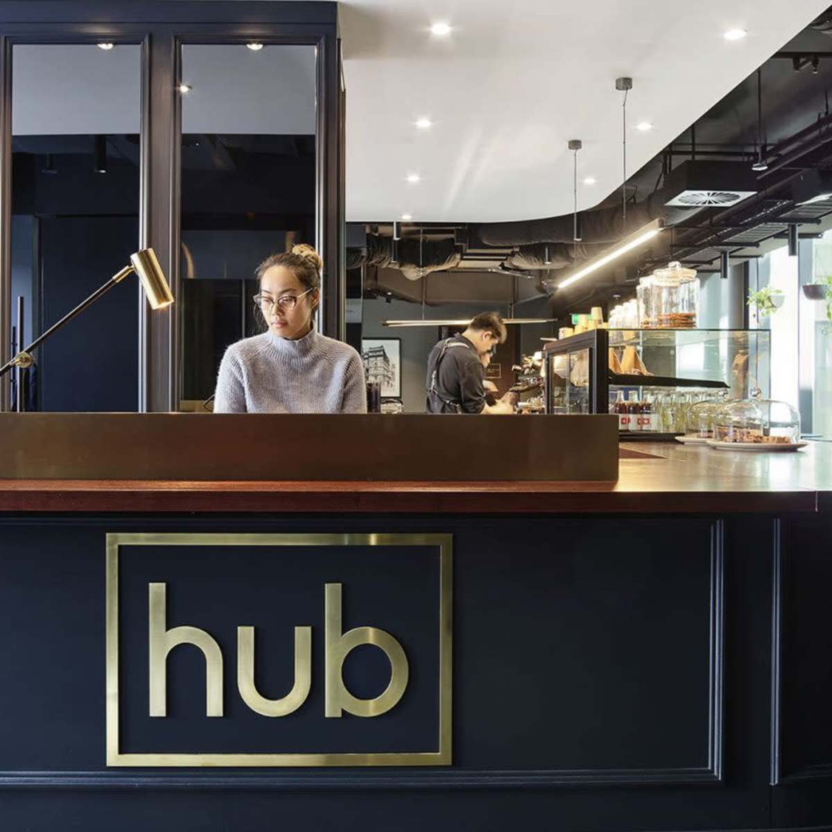 A modern reception area features a dark counter labeled "hub," with a desk attendant and a cafe space in the background.