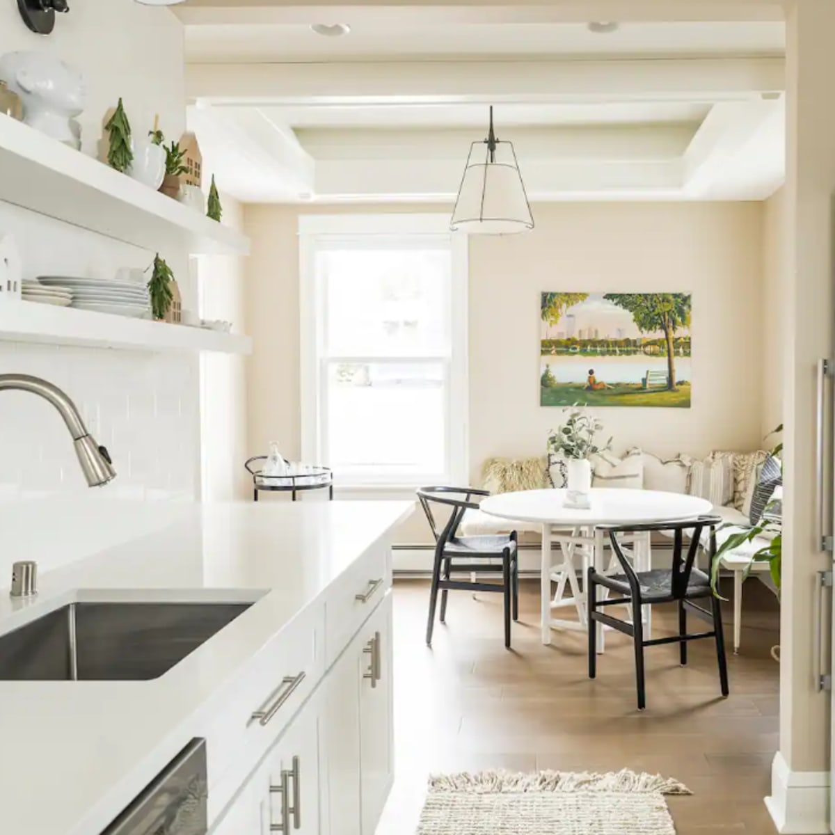 A bright kitchen features white cabinetry and a sleek sink, leading into a dining area with a round table and a colorful painting on the wall.