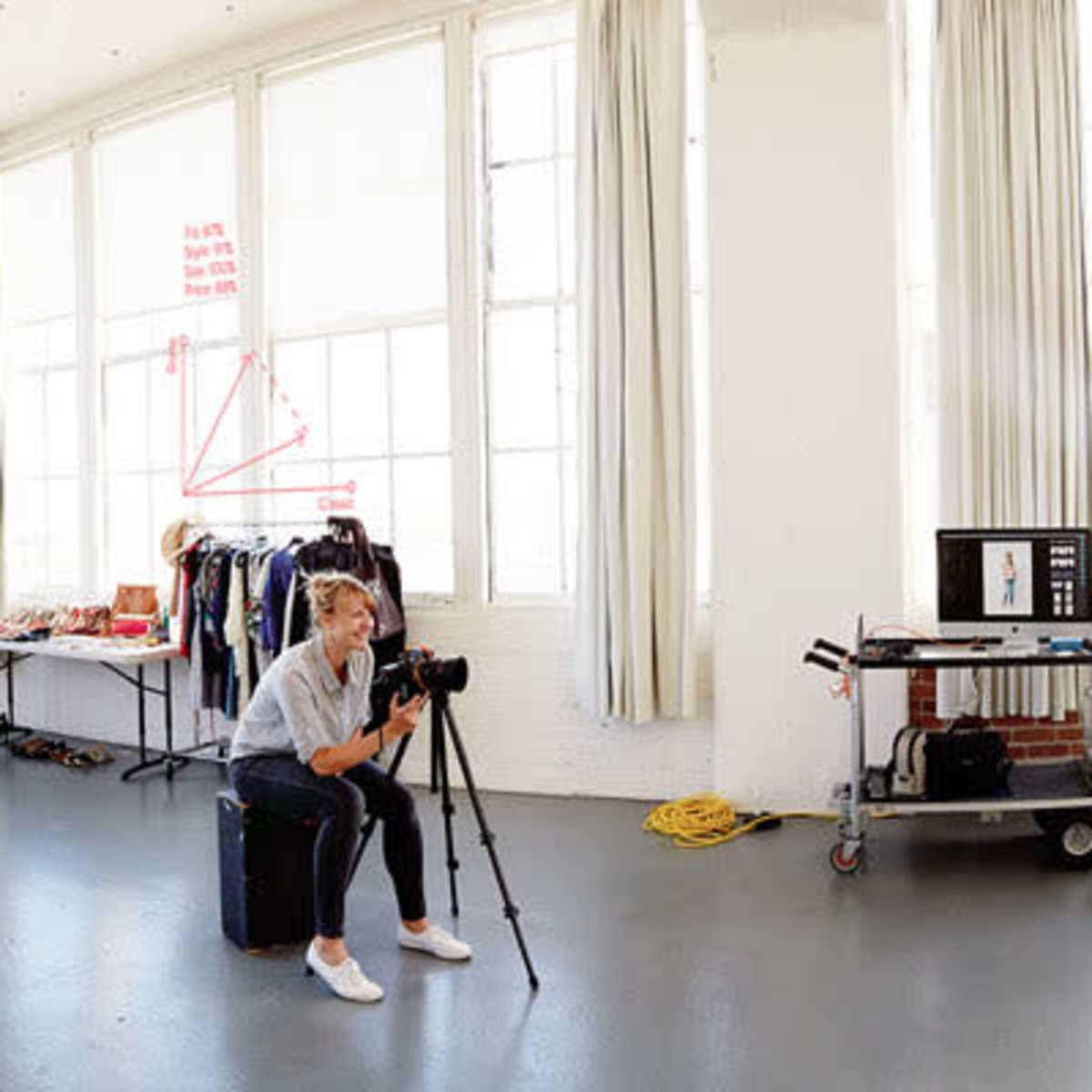 A photographer captures a model in a brightly lit studio with large windows and a white backdrop featuring diagrams and equations.