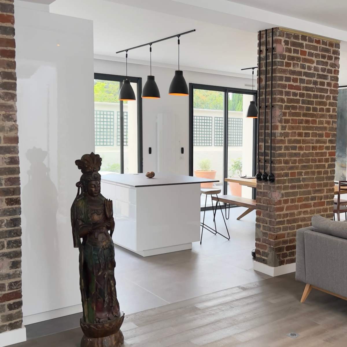 A modern living space with a gray sofa, a decorative statue, exposed brick columns, and an open kitchen area with pendant lighting.