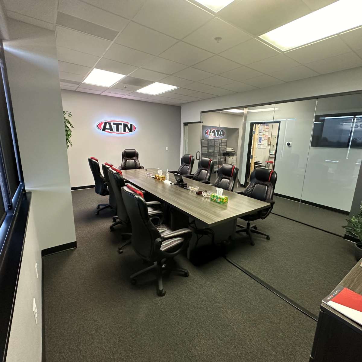 A modern office meeting room with a long conference table surrounded by black ergonomic chairs and glass walls.