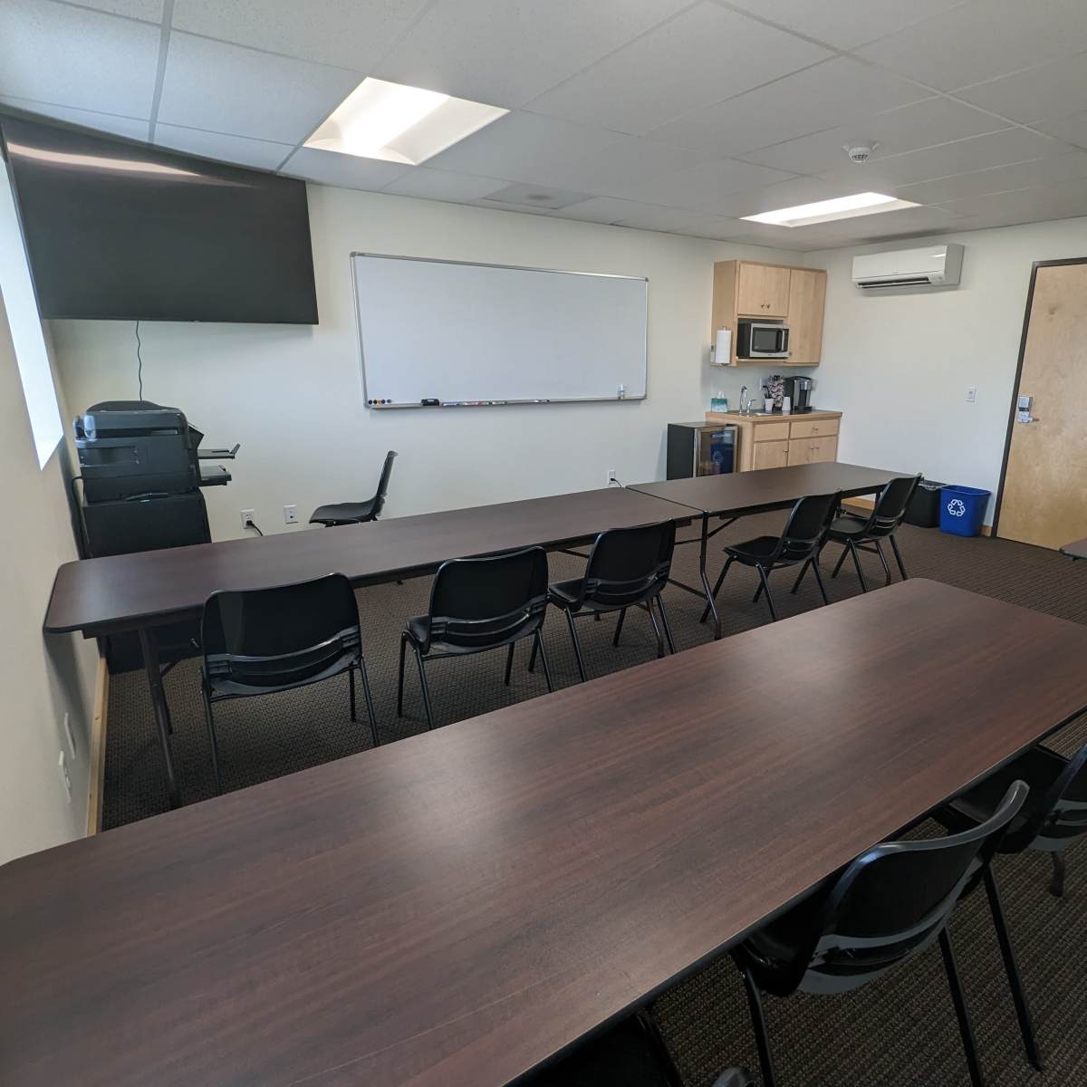 A conference room with several tables arranged in a U-shape, chairs placed around them, a whiteboard, a kitchenette area, and a television mounted on the wall.