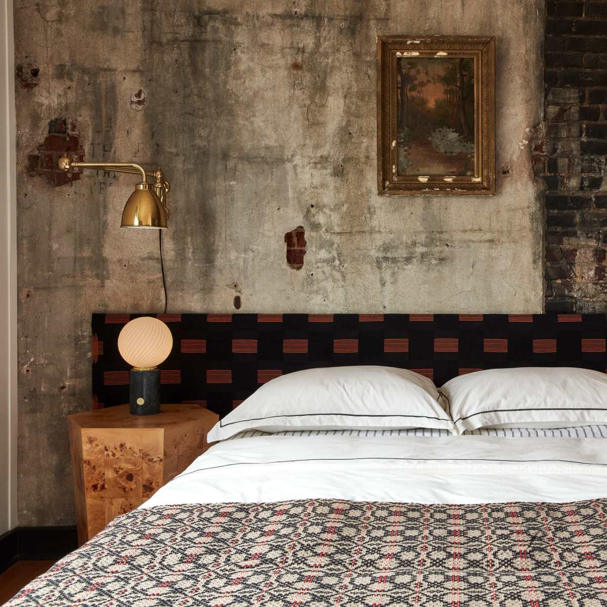 A bed with a patterned blanket against a textured wall featuring exposed brick and concrete, flanked by two bedside tables with lamps.