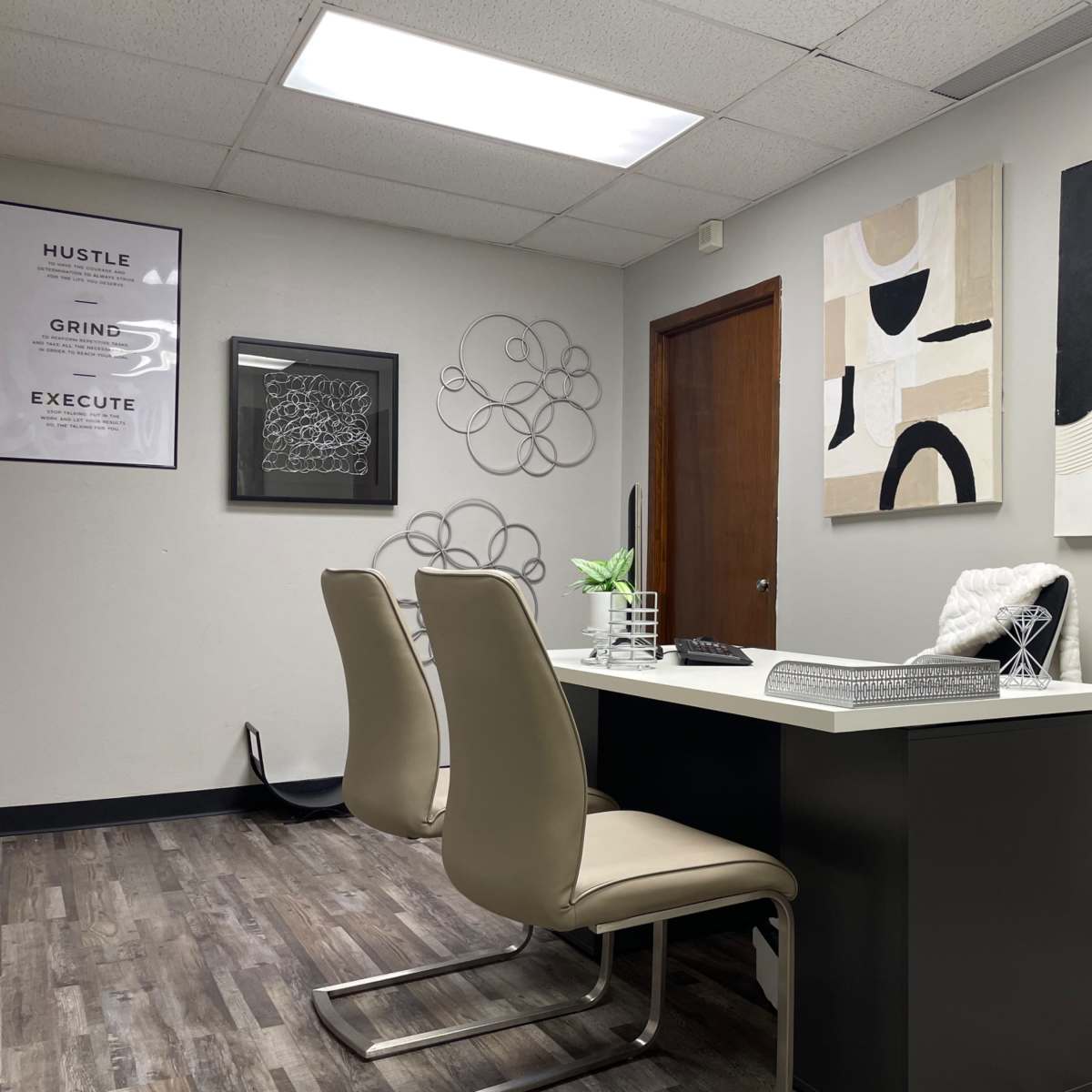 A modern office space featuring a desk with two chairs, decorative wall art, and a closed wooden door in the background.