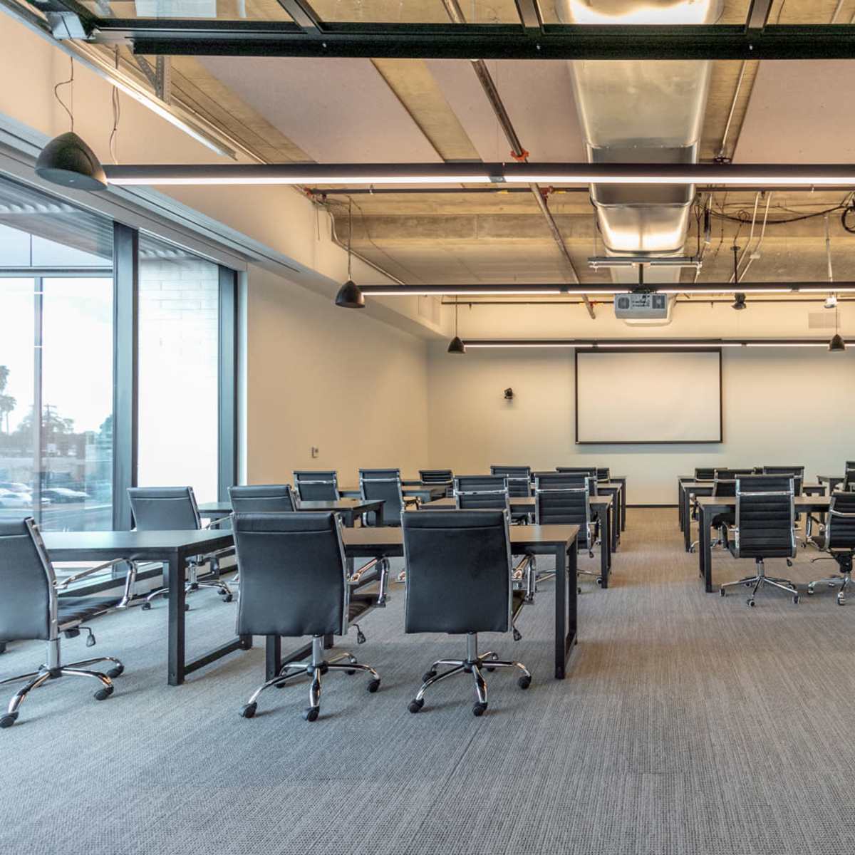 A conference room is set up with multiple black chairs and tables arranged for a meeting, featuring a projector on the ceiling and a blank screen on the wall.