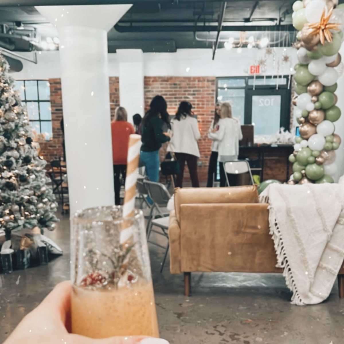 A person holds a glass of orange beverage with a striped straw while standing in a decorated event space featuring a Christmas tree and balloons.