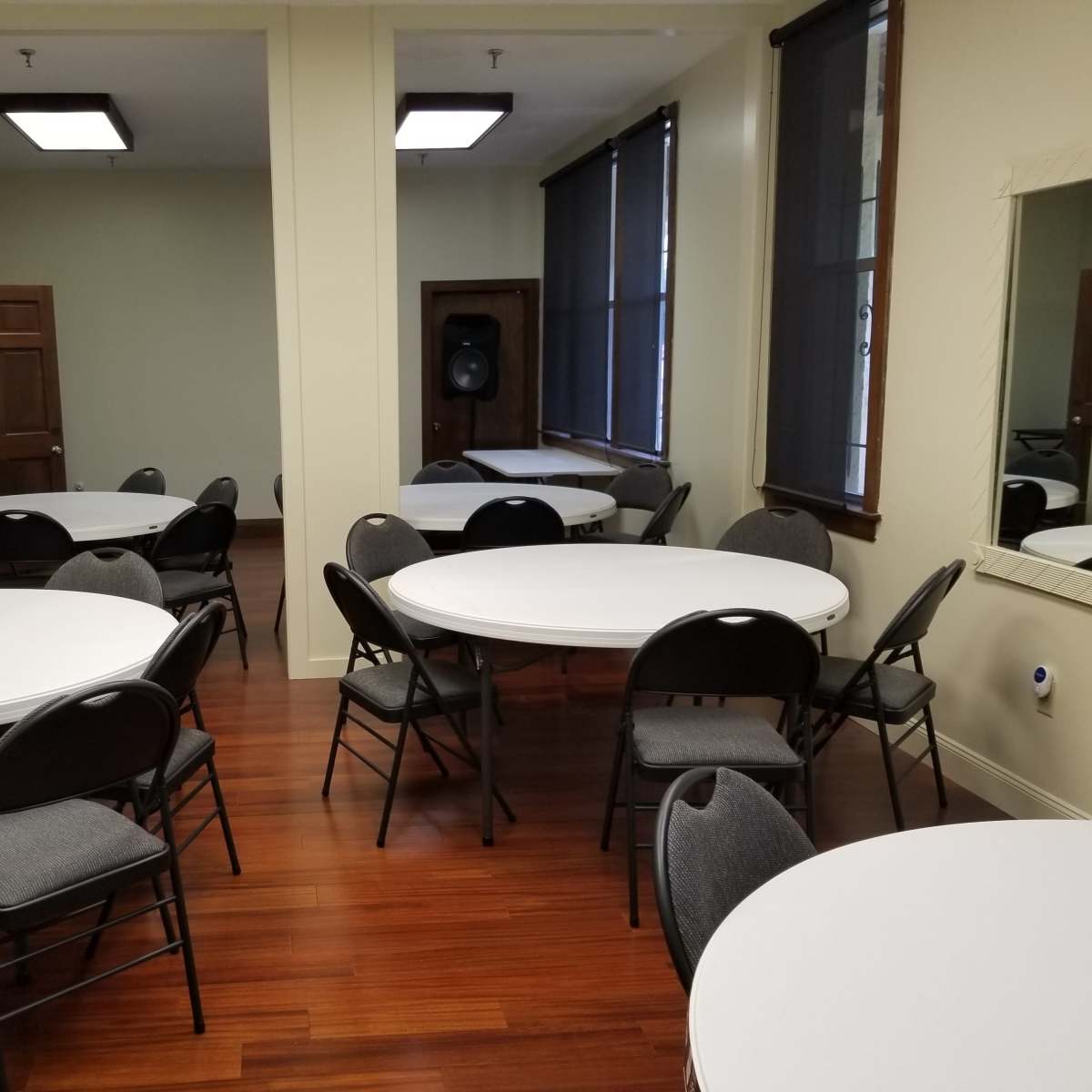 A room with several circular tables surrounded by chairs, featuring wooden floors and light-colored walls.