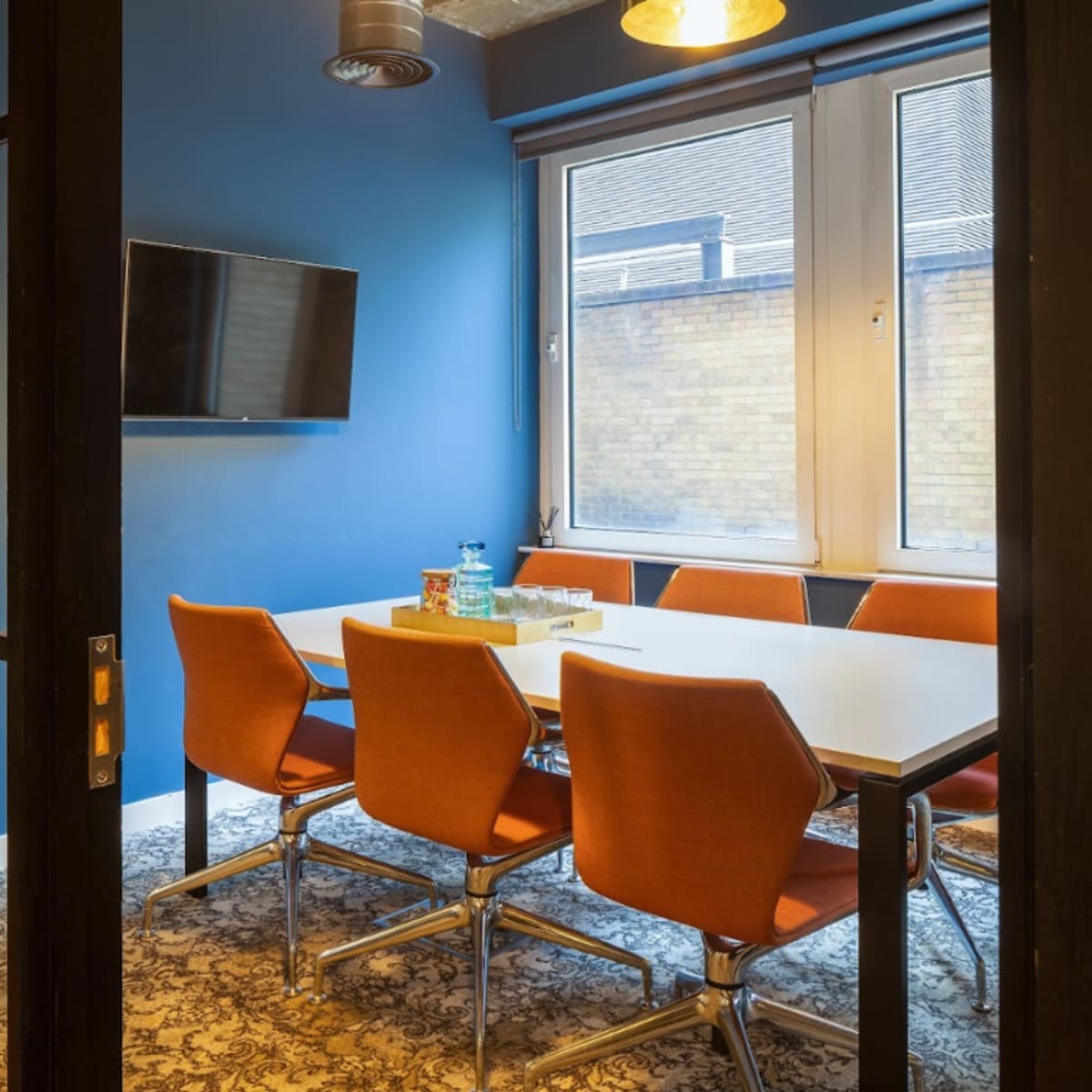 A meeting room features a large table surrounded by orange chairs, with a television mounted on the wall and large windows allowing natural light.