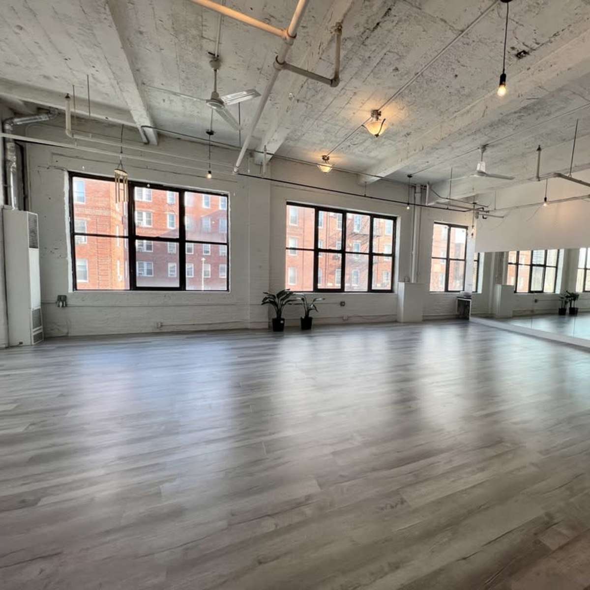 A spacious, well-lit studio with large windows and polished wooden flooring, featuring a mirror along one wall.