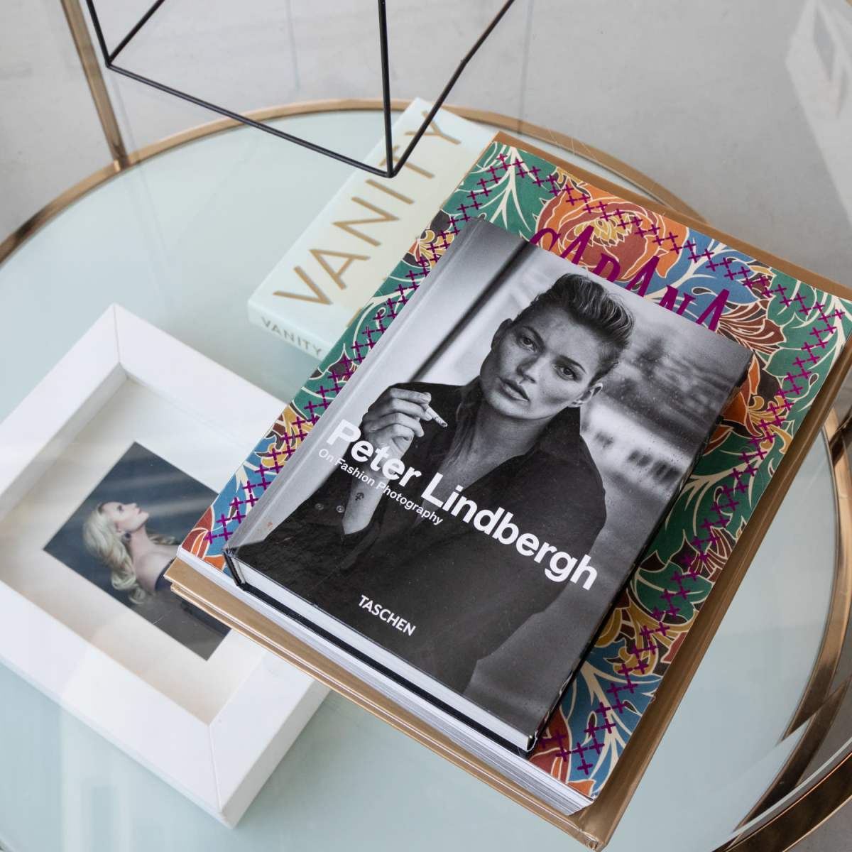 A glass table displays several books, including a photograph book by Peter Lindbergh, alongside a framed picture and a white box labeled "Vanity."