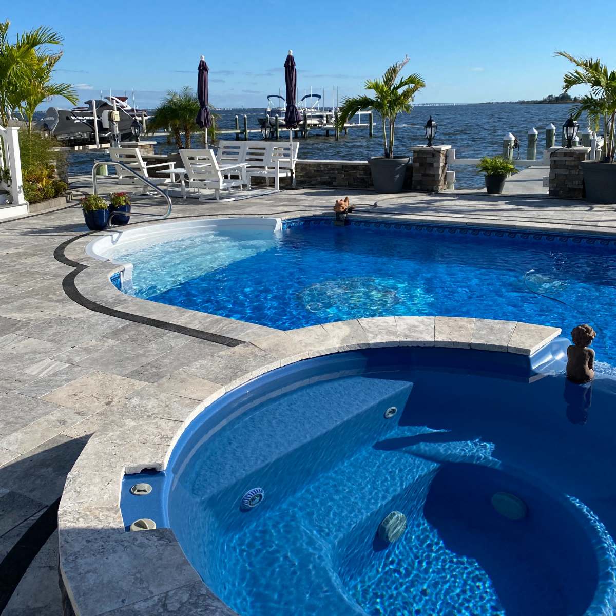 A swimming pool area with a spa, surrounded by stone paving and overlooking a body of water with boats in the background.