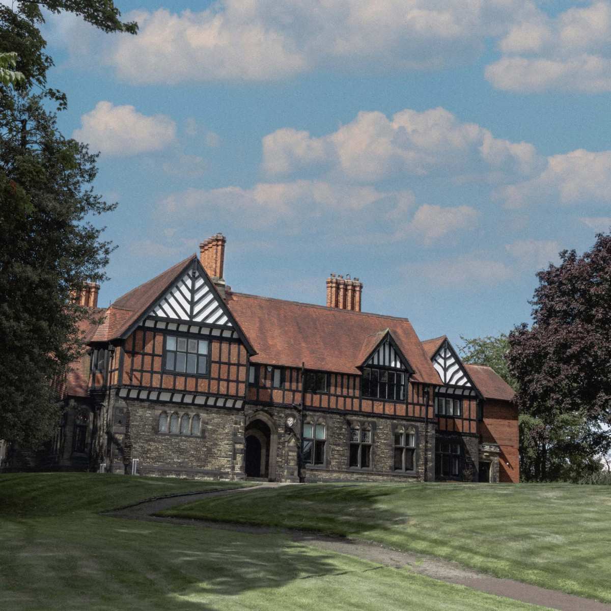 A large, two-story Tudor-style mansion with a red tiled roof is situated on a grassy hill surrounded by trees under a partly cloudy sky.