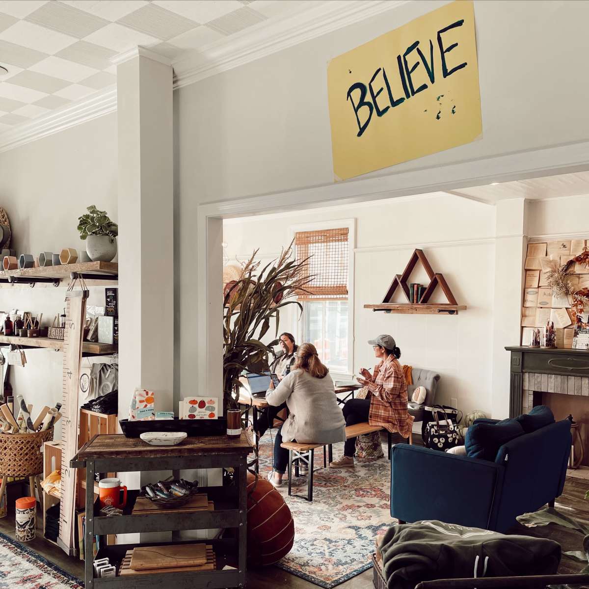 A cozy room features a group of three people interacting around a table, with various decorative items and plants visible throughout the space.