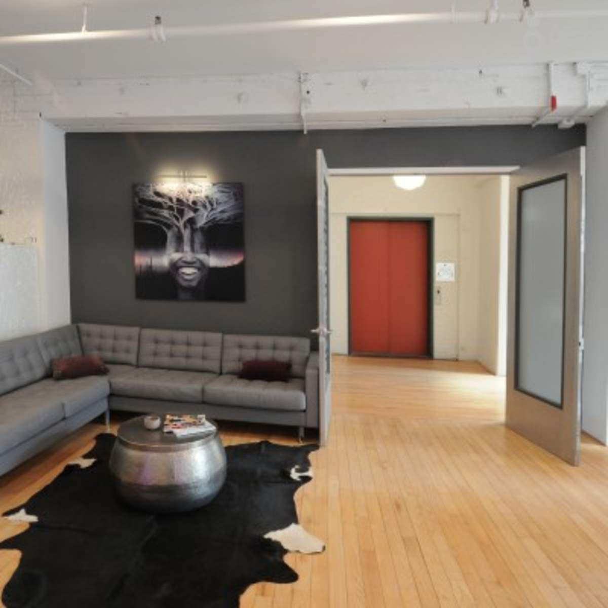 A modern lounge area with a sectional gray sofa, a round metallic coffee table, a black and white artwork on the wall, and an entrance leading to an elevator in the background.