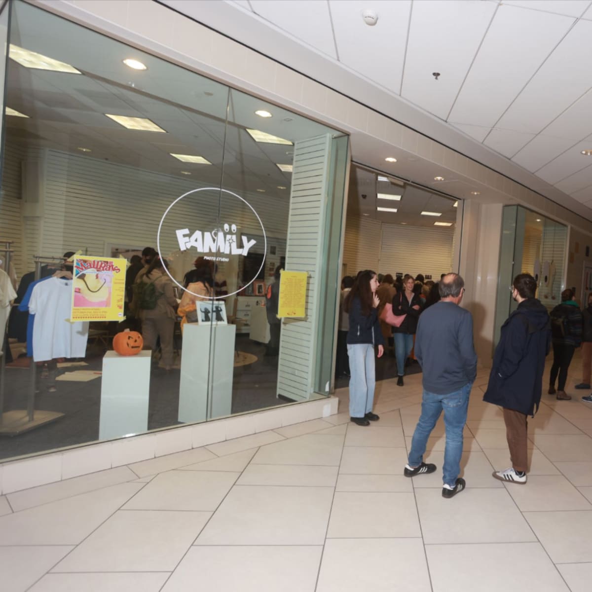 A group of people gathers in a shopping mall near a store called "FAMILY," with windows displaying clothing and decorations.