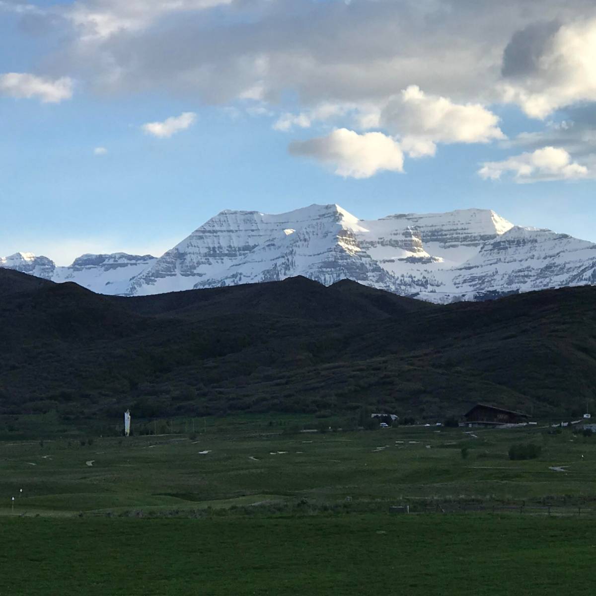 A mountainous landscape with snow-capped peaks rising above green hills and a valley, under a partly cloudy sky.