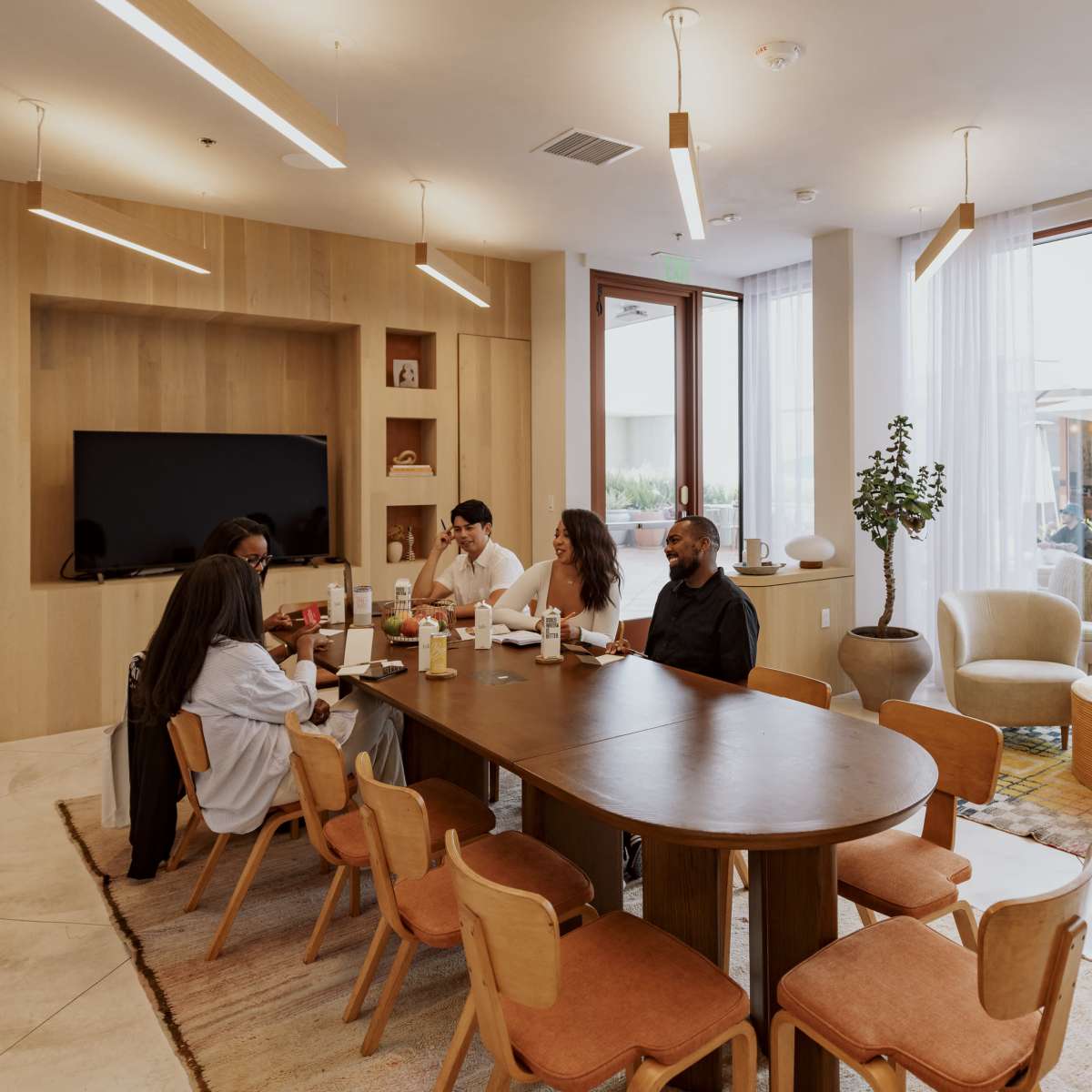 A group of five people is sitting around a wooden table in a modern, well-lit common area with large windows and comfortable seating.
