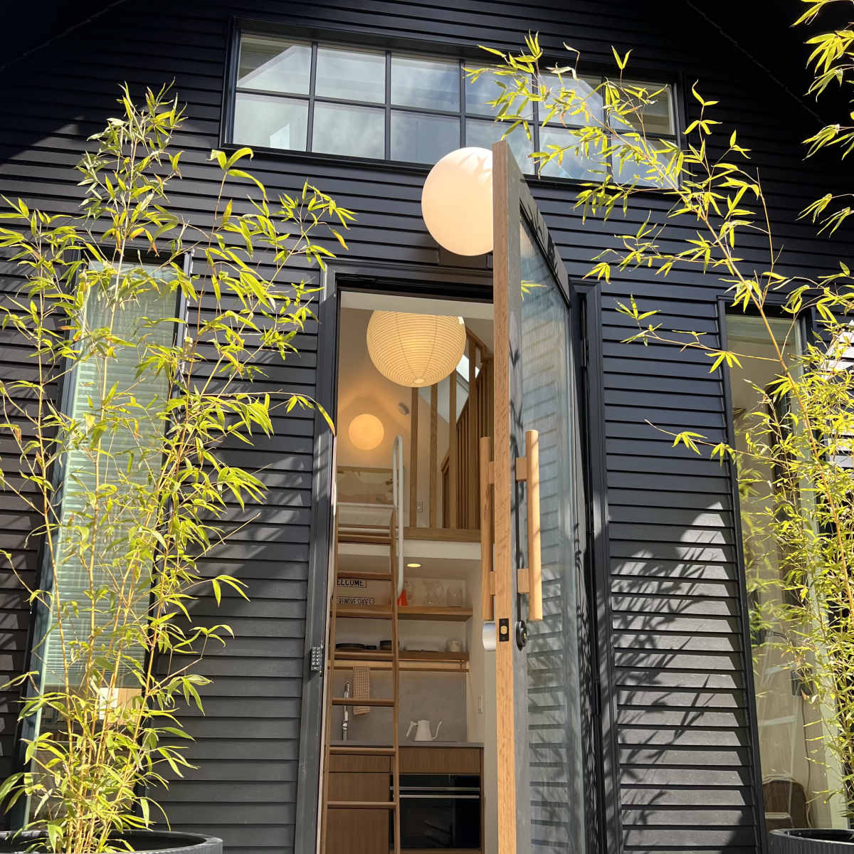 A modern black house features an open front door flanked by tall bamboo plants in planters, leading into a bright interior with a visible ladder and round pendant lights.