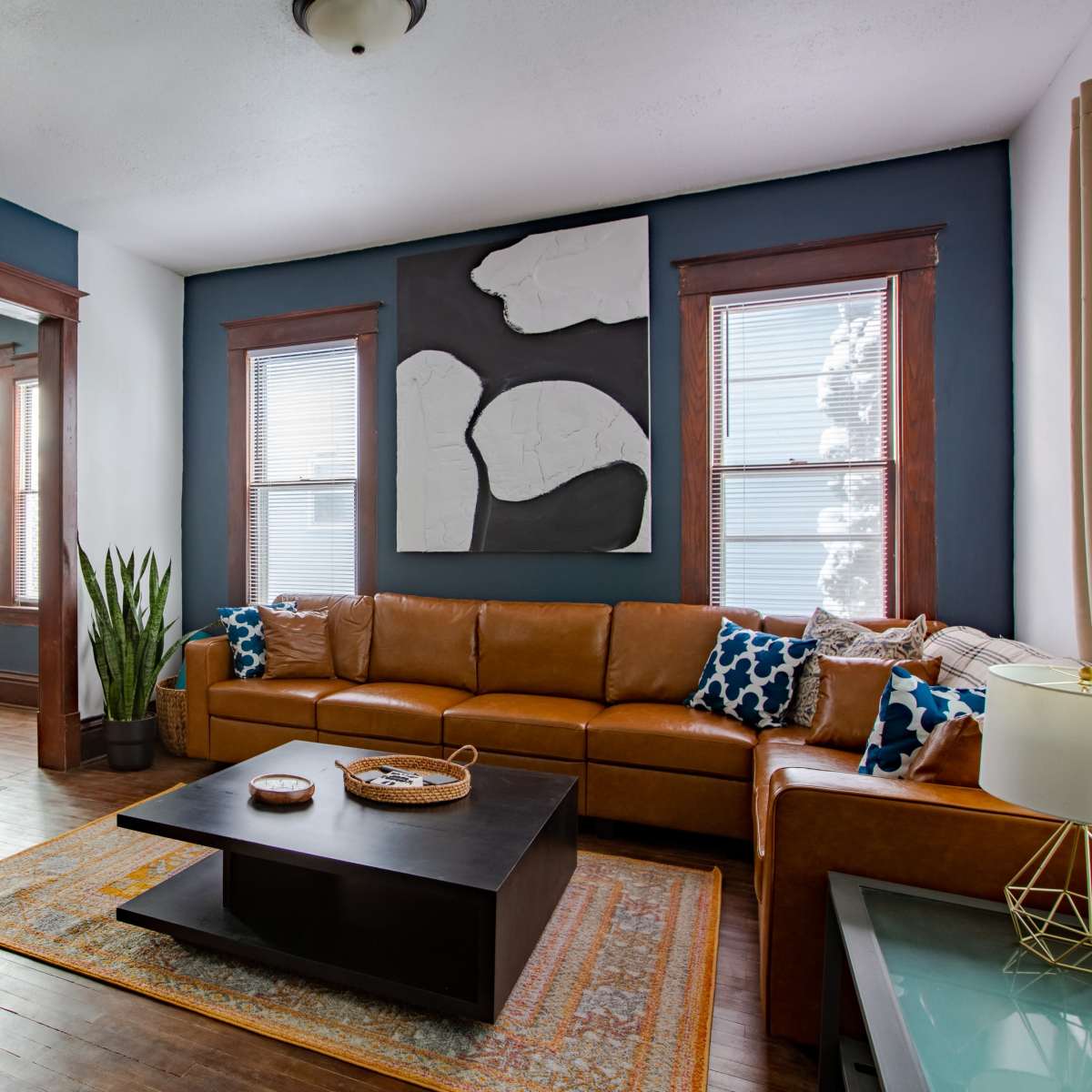 A living room with a brown leather sectional sofa, a black coffee table, and a large abstract wall decoration, with a dining area visible through an archway.