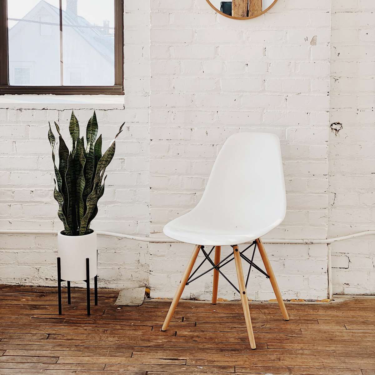 A minimalist interior featuring a white chair, a round mirror mounted on a white brick wall, and a potted plant in a black and white planter.