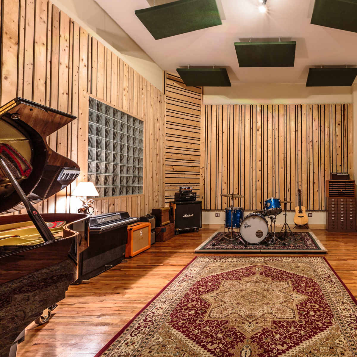 A spacious recording studio with wooden walls, a drum set, electric guitars, and various musical equipment arranged around an ornate area rug.