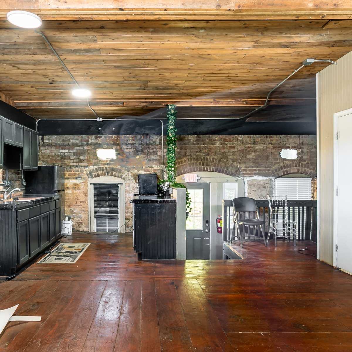 A rustic kitchen area with wooden floors, exposed brick walls, and black cabinetry, leading to a dining space with chairs.