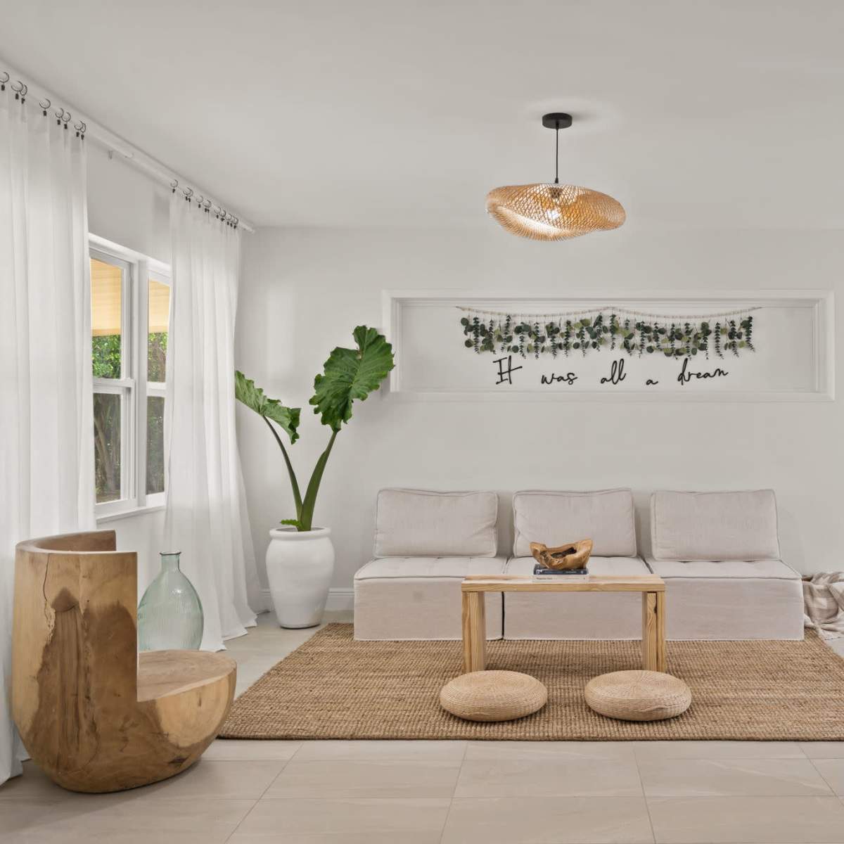 A living room featuring a light-colored couch, a wooden coffee table, two circular ottomans, and decor elements like a large plant and wall artwork.
