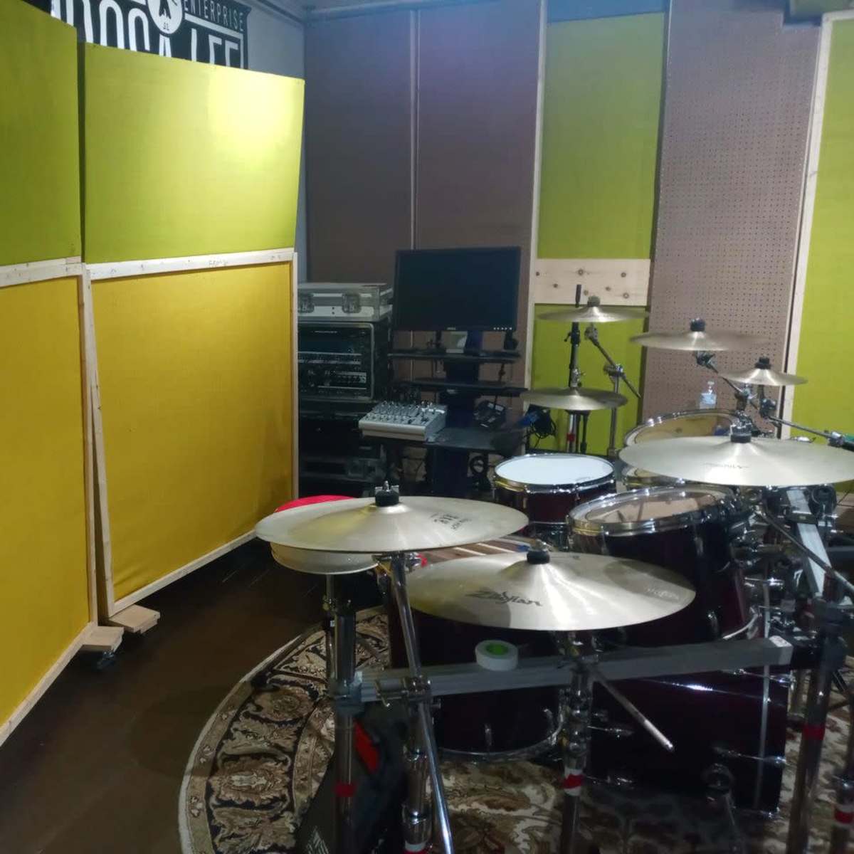 A section of a music studio featuring a drum set, large green acoustic panels, and various audio equipment in the background.