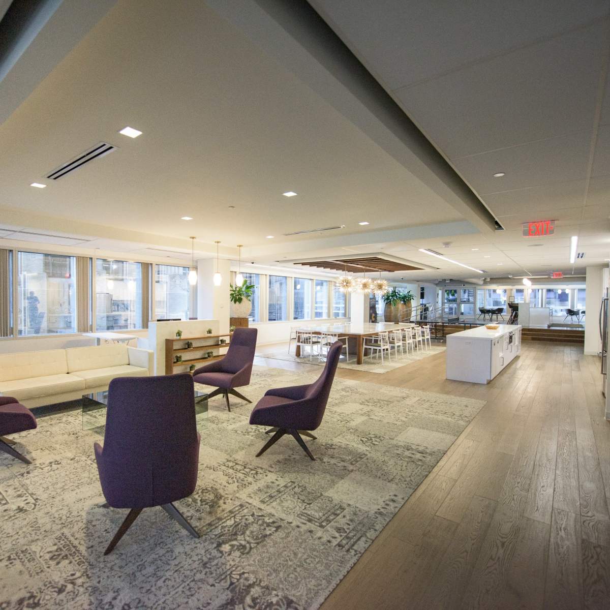 A modern office space with seating areas including purple chairs, a white sofa, and dining tables, illuminated by natural light from large windows.