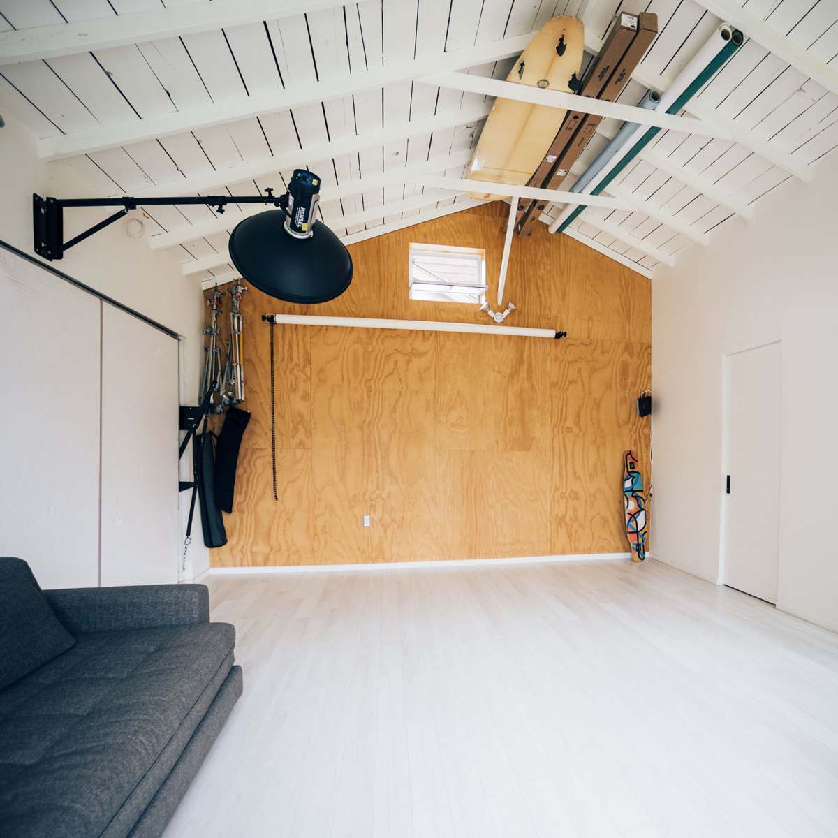 A spacious, minimally furnished room with a wooden wall, a gray sofa, and a desk against a white interior.
