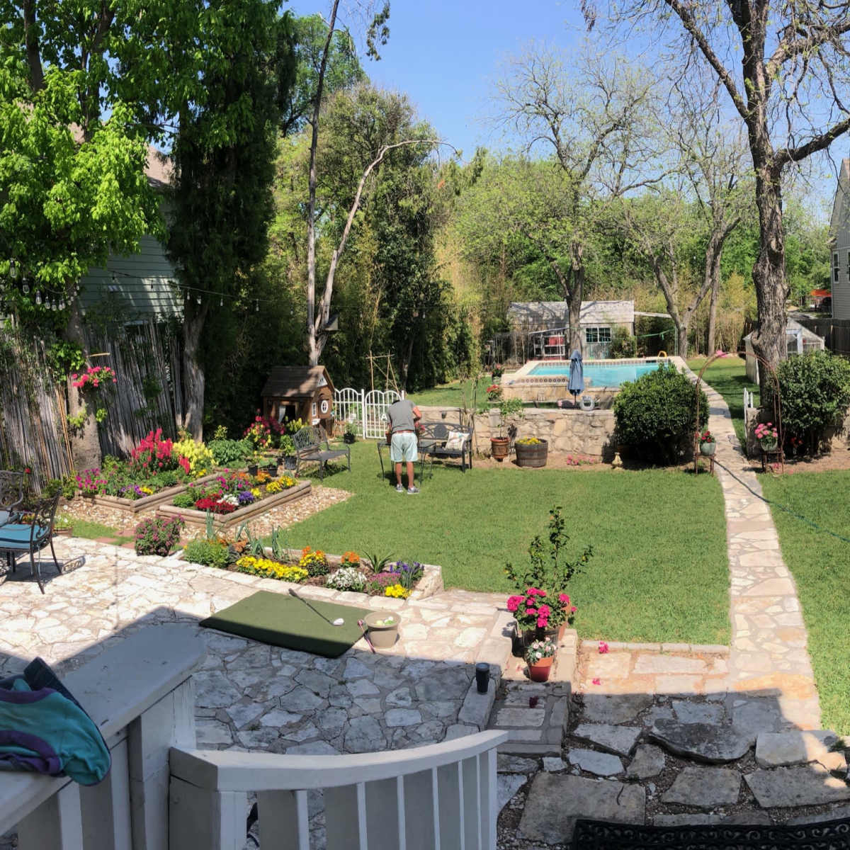 A backyard scene features a grassy area with a swimming pool, stone landscaping, flower beds, and a person tending to the garden.