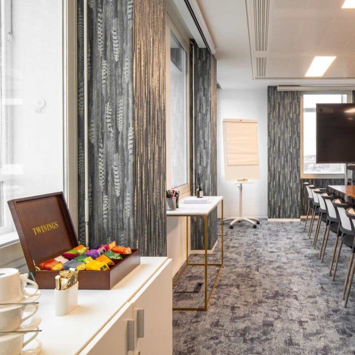 A modern conference room features a long wooden table set with glass water bottles, a large screen at one end, and a flip chart on a stand.